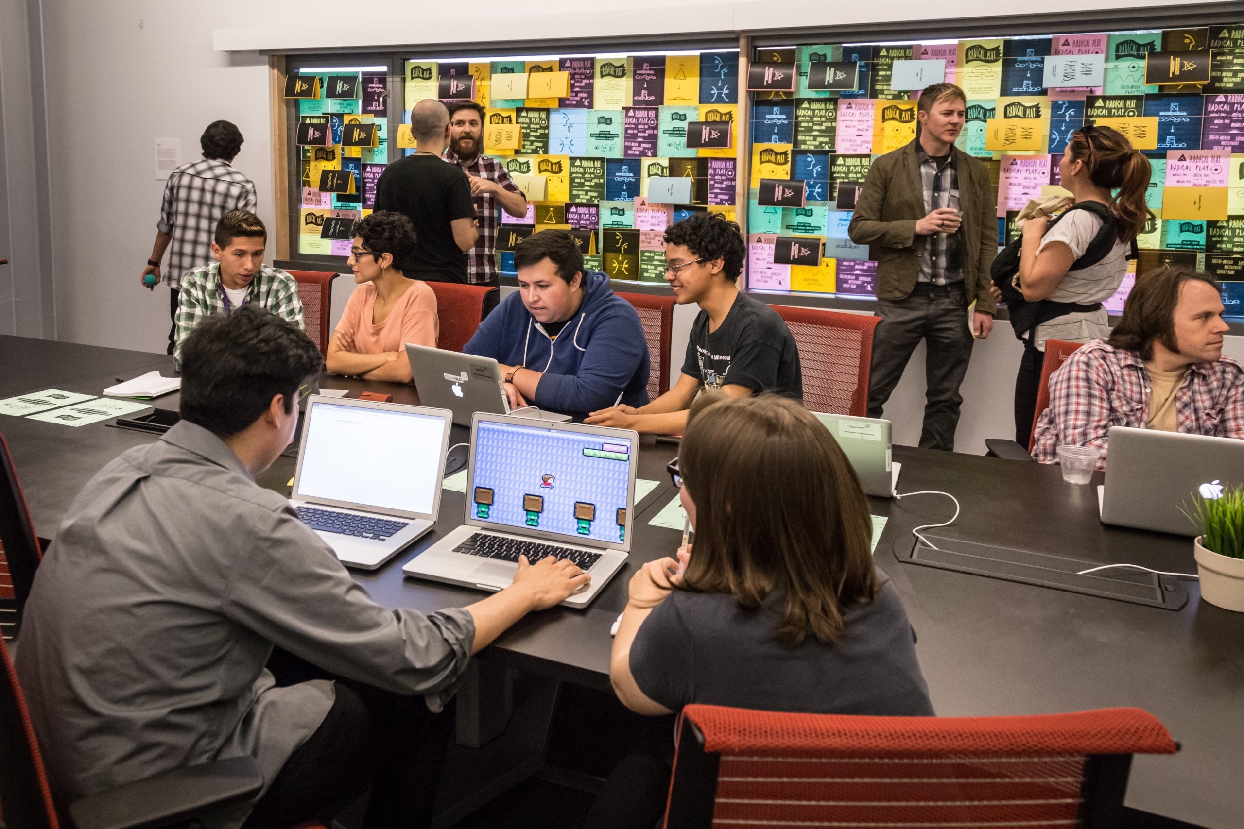 Young adults collaborating on laptops and notes around a table in a modern classroom or co-working space, with a wall of colorful sticky notes in the background.