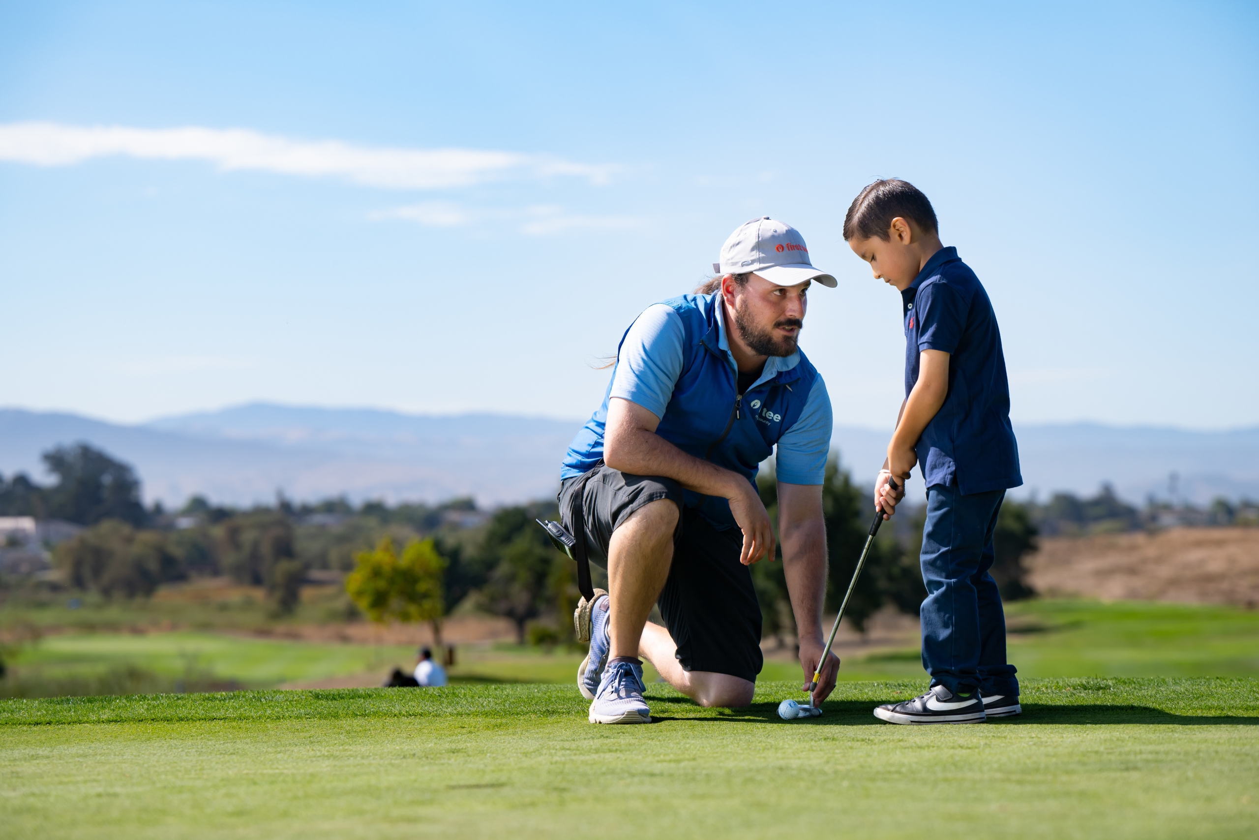A golf instructor kneeling on the green to help a young boy practice his putting swing, with hills and mountains in the background.