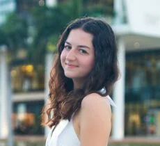 Headshot of a smiling young woman with dark hair, looking over her shoulder with an urban building backdrop.