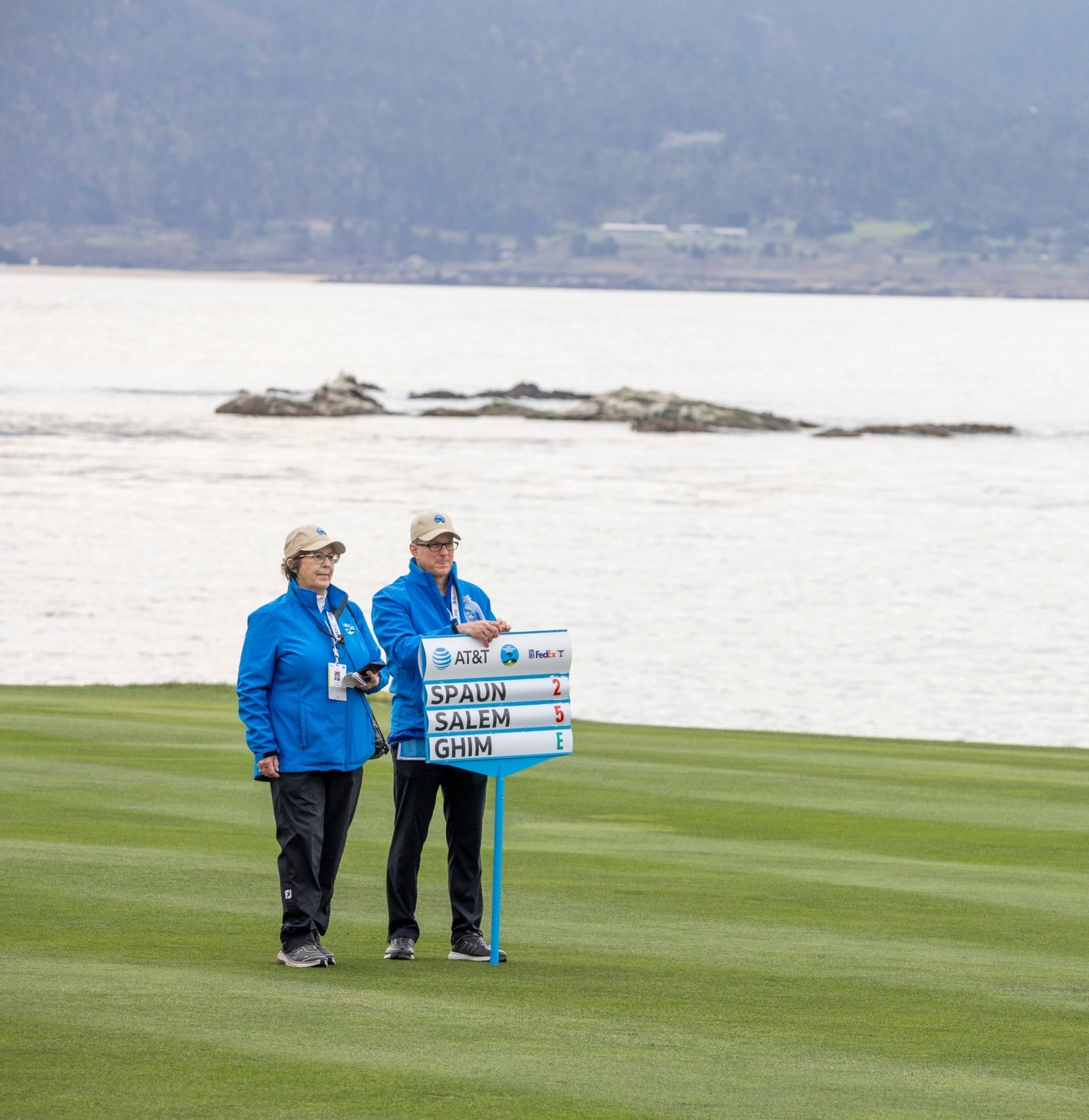 Two tournament volunteers in blue jackets holding a large manual golf scoreboard on the green overlooking the ocean.