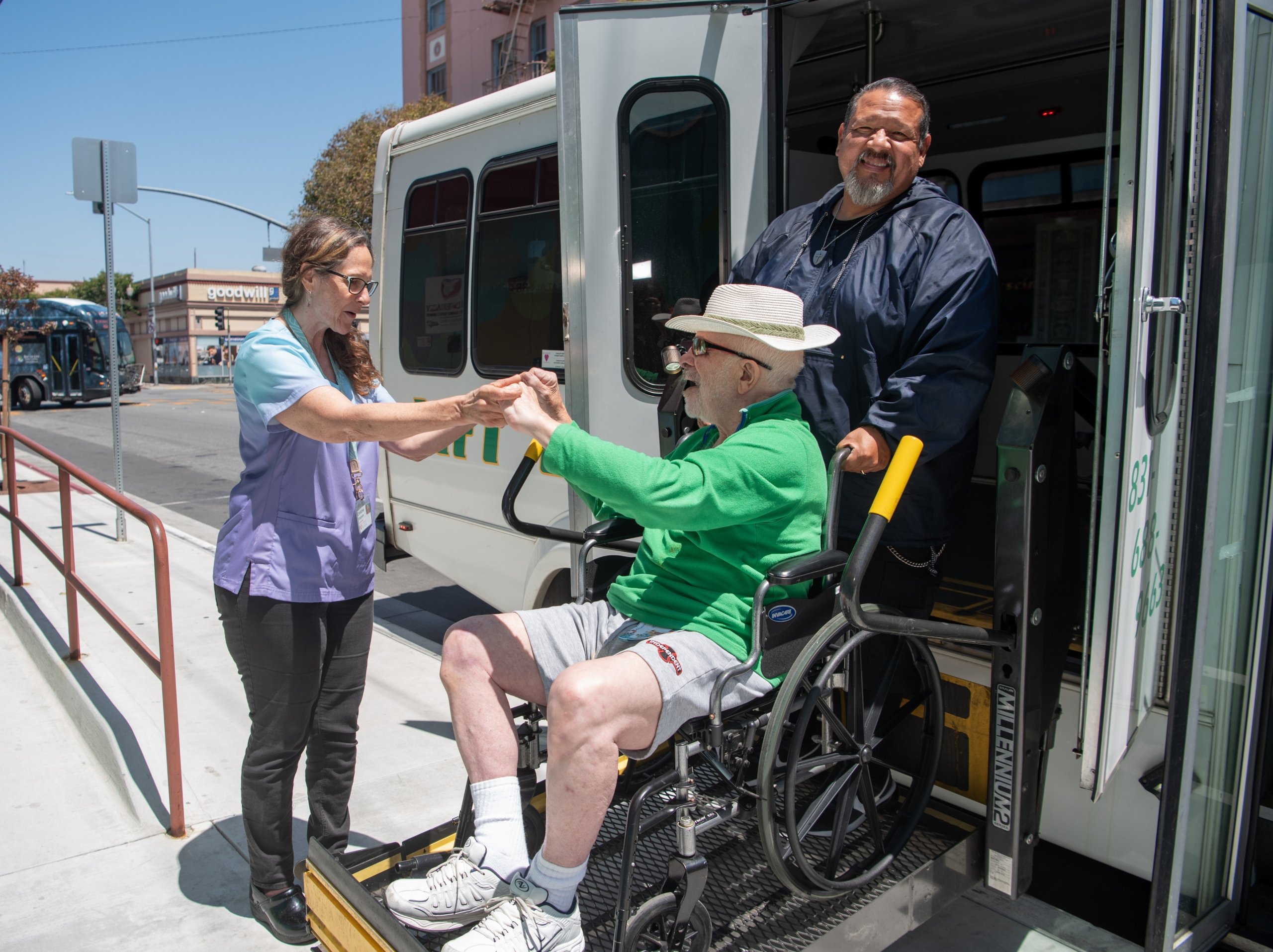 A driver and a female caregiver help a man in a wheelchair off a transit van using a hydraulic lift in an urban setting.