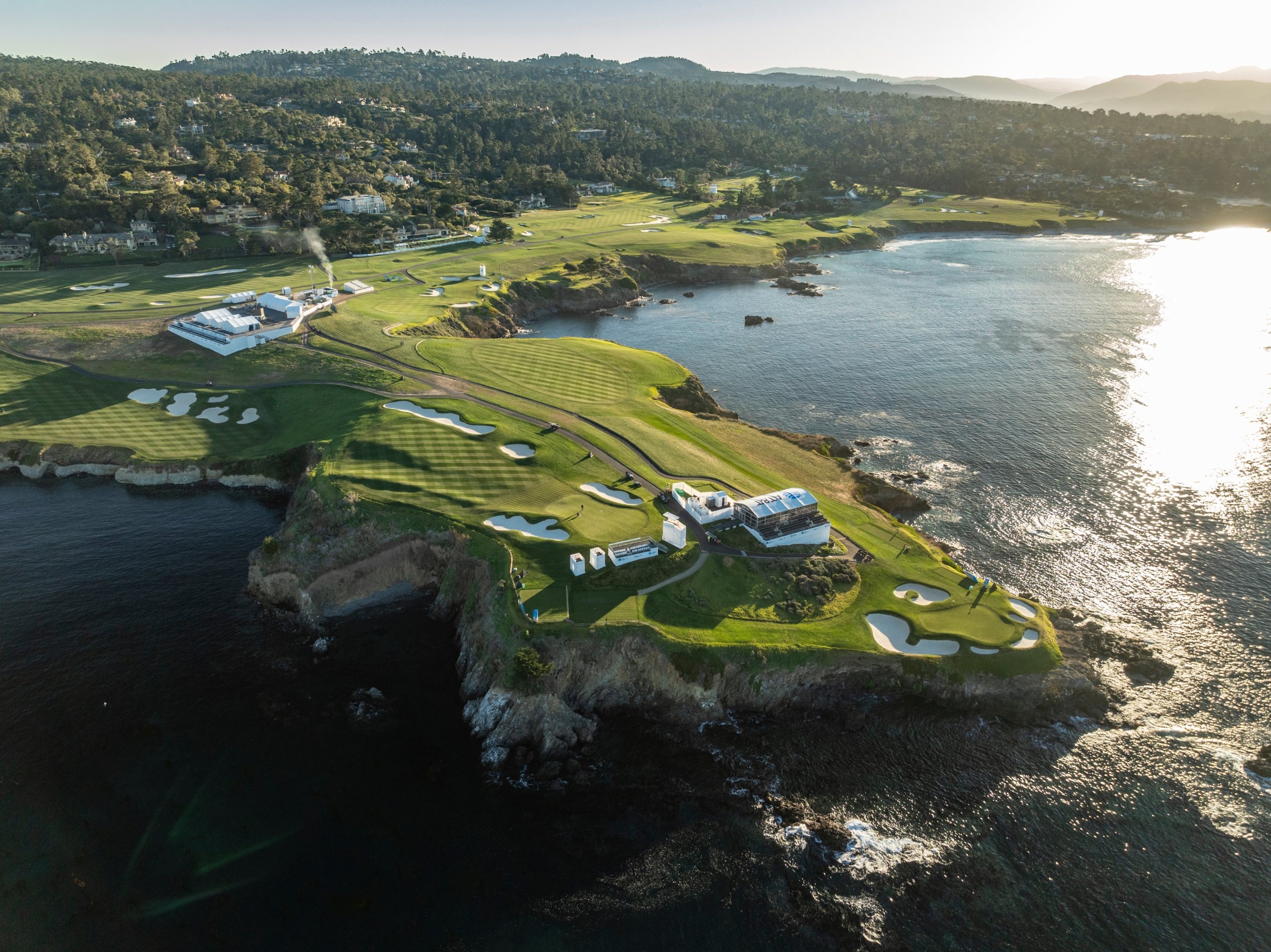 An aerial view of a dramatic, seaside golf course peninsula featuring greens, bunkers, and event structures overlooking the Pacific Ocean.