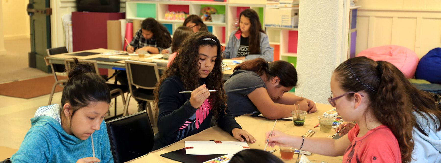 Female students concentrating on painting and creative activities at tables in a brightly lit community art classroom.