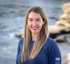 Headshot of a smiling woman with long blonde hair wearing a blue sweater, standing near the ocean with rocky shoreline visible.