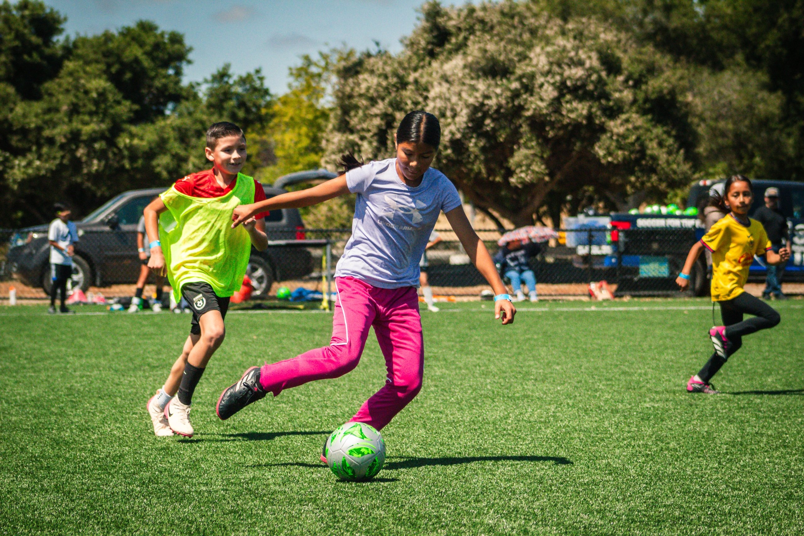 Three children running and playing soccer vigorously on a green turf field during a youth sports program.