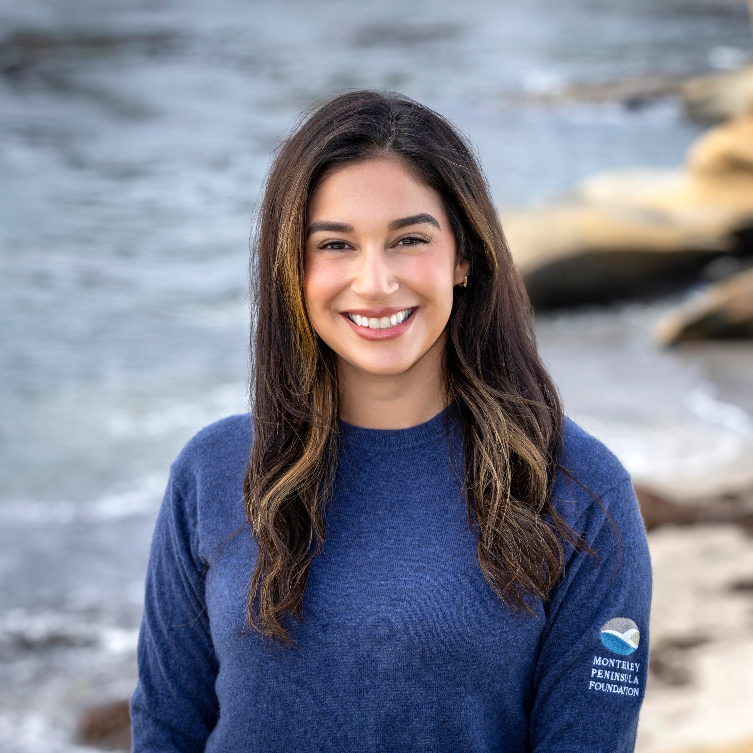 Headshot of a smiling woman with long brown hair wearing a blue MPF branded sweater, set against a blurred coastal background.