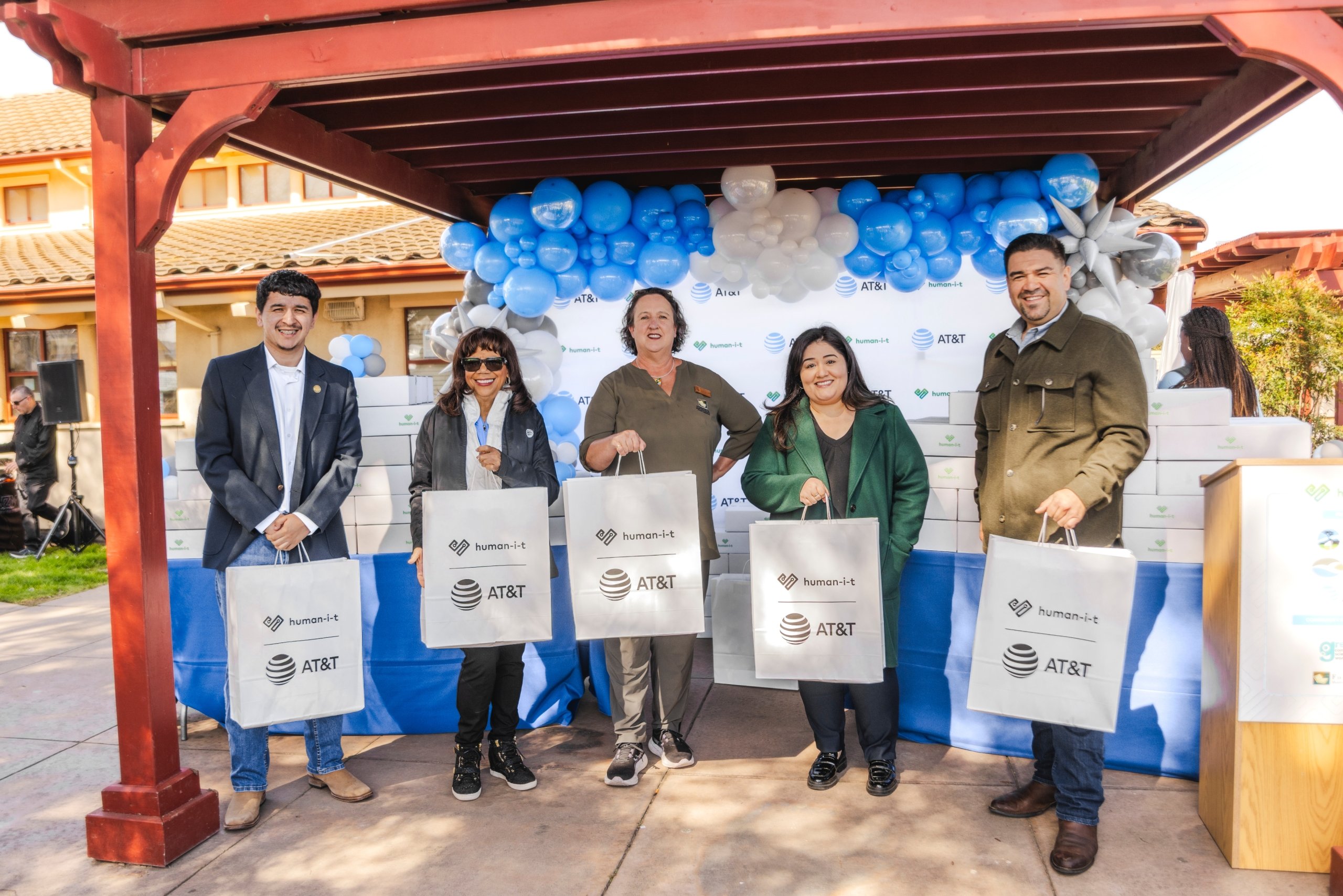 Five individuals, including representatives from human-I-T and AT&T, posing under an arbor with bags of tech during a community giveaway event.