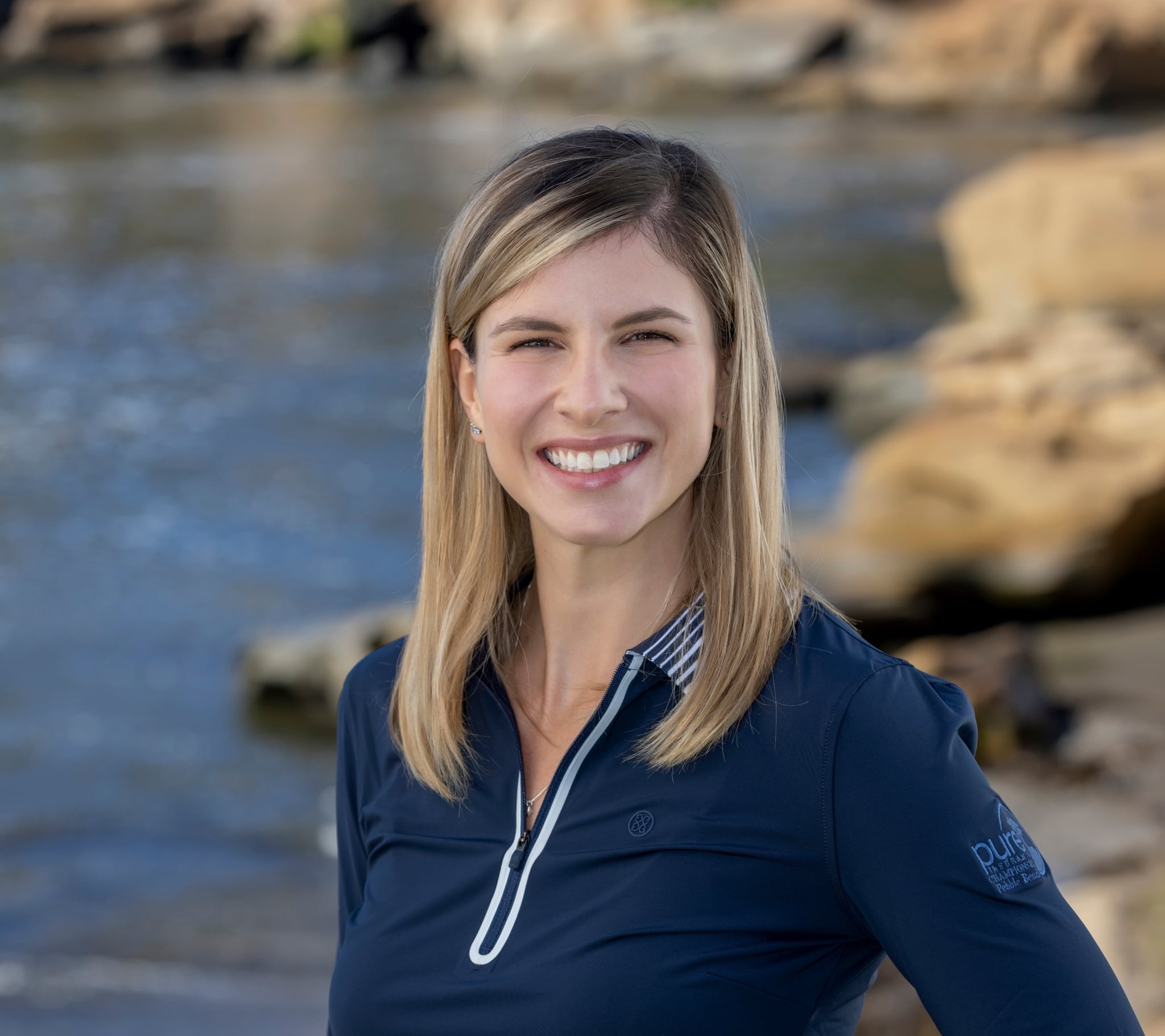 Headshot of a smiling woman with blonde hair wearing a navy blue collared golf shirt, standing by the water.