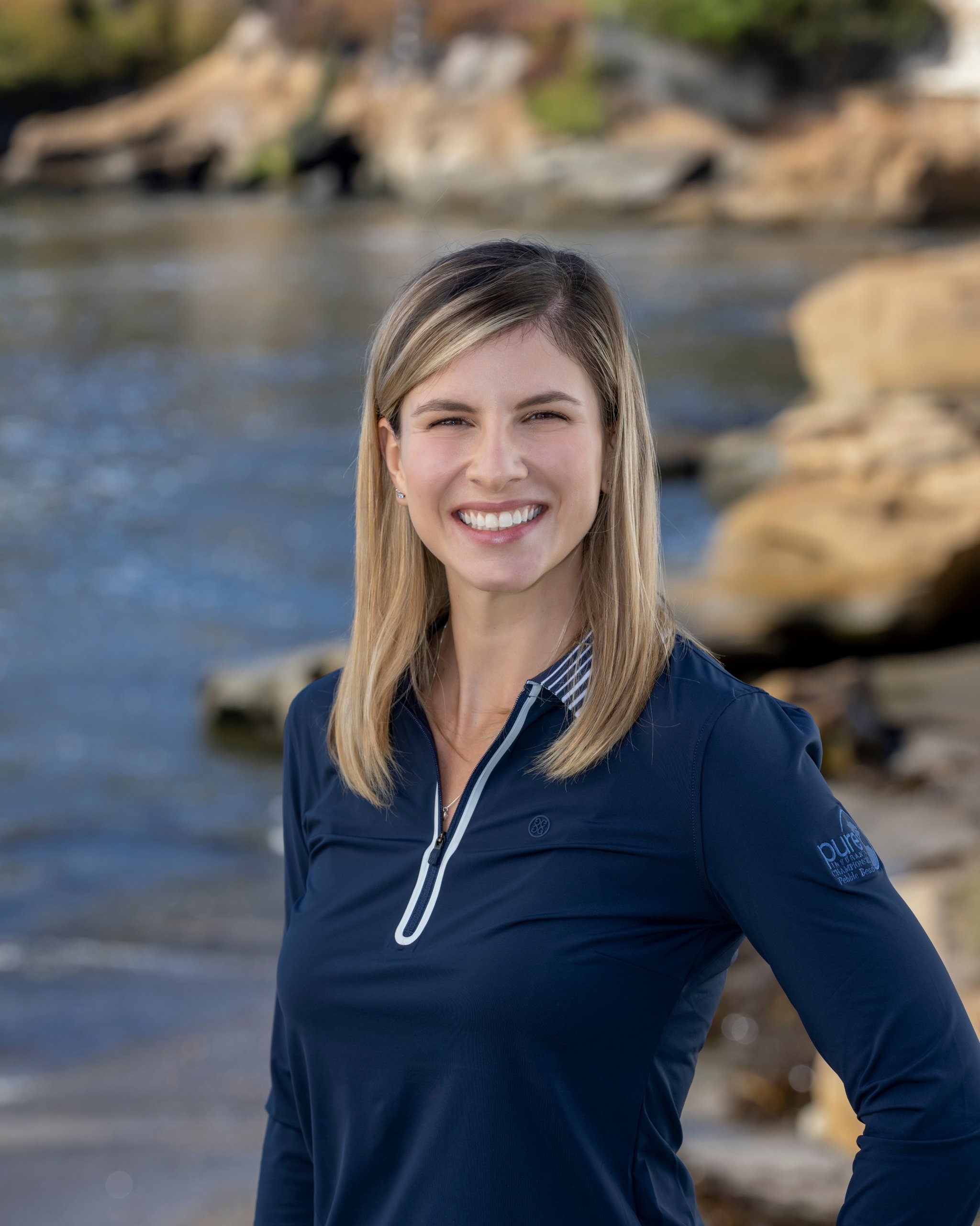 Headshot of a smiling woman with blonde hair wearing a navy blue collared golf shirt, standing by the water.