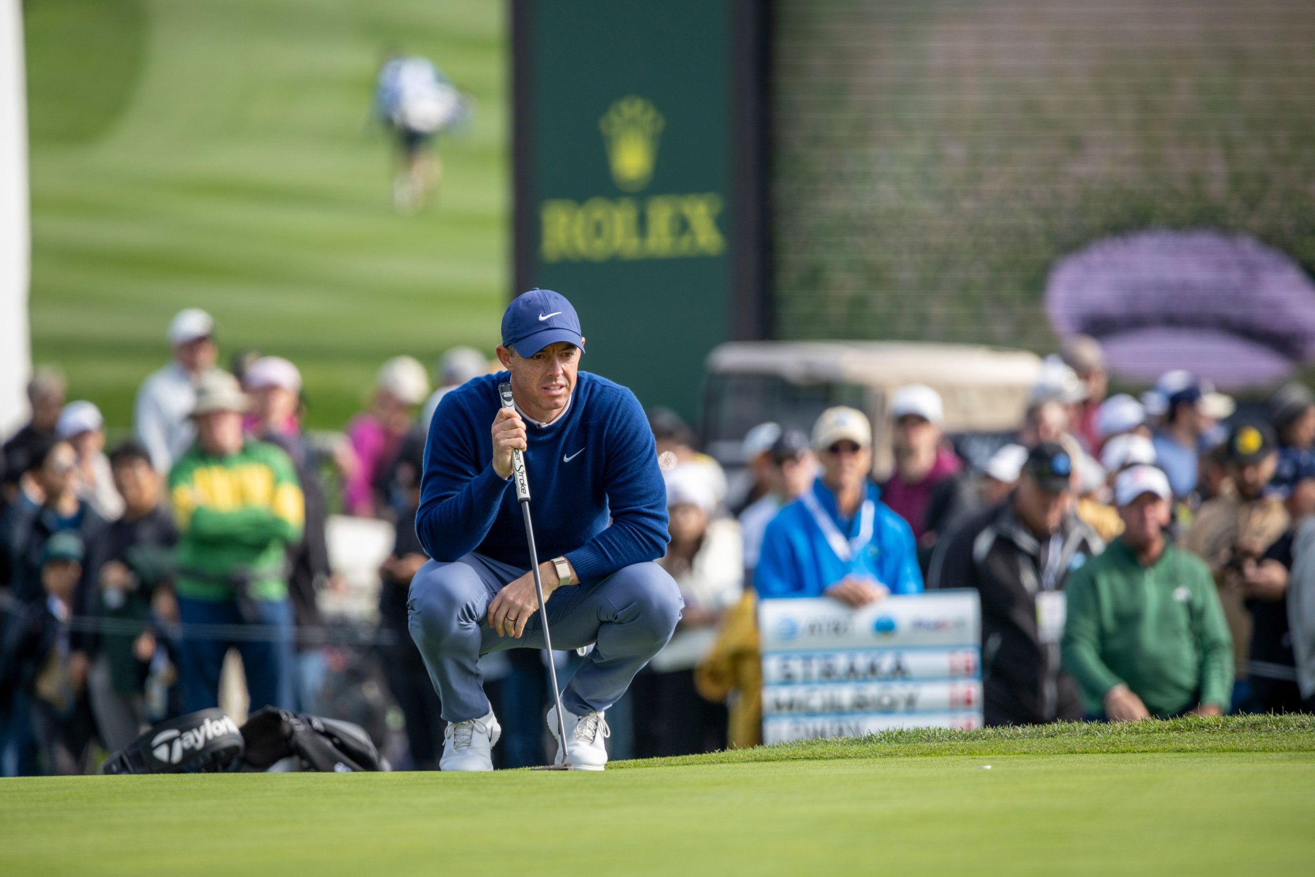 Golfer Rory McIlroy crouching on the green to line up a putt during the tournament, with a crowd and Rolex banner in the background.
