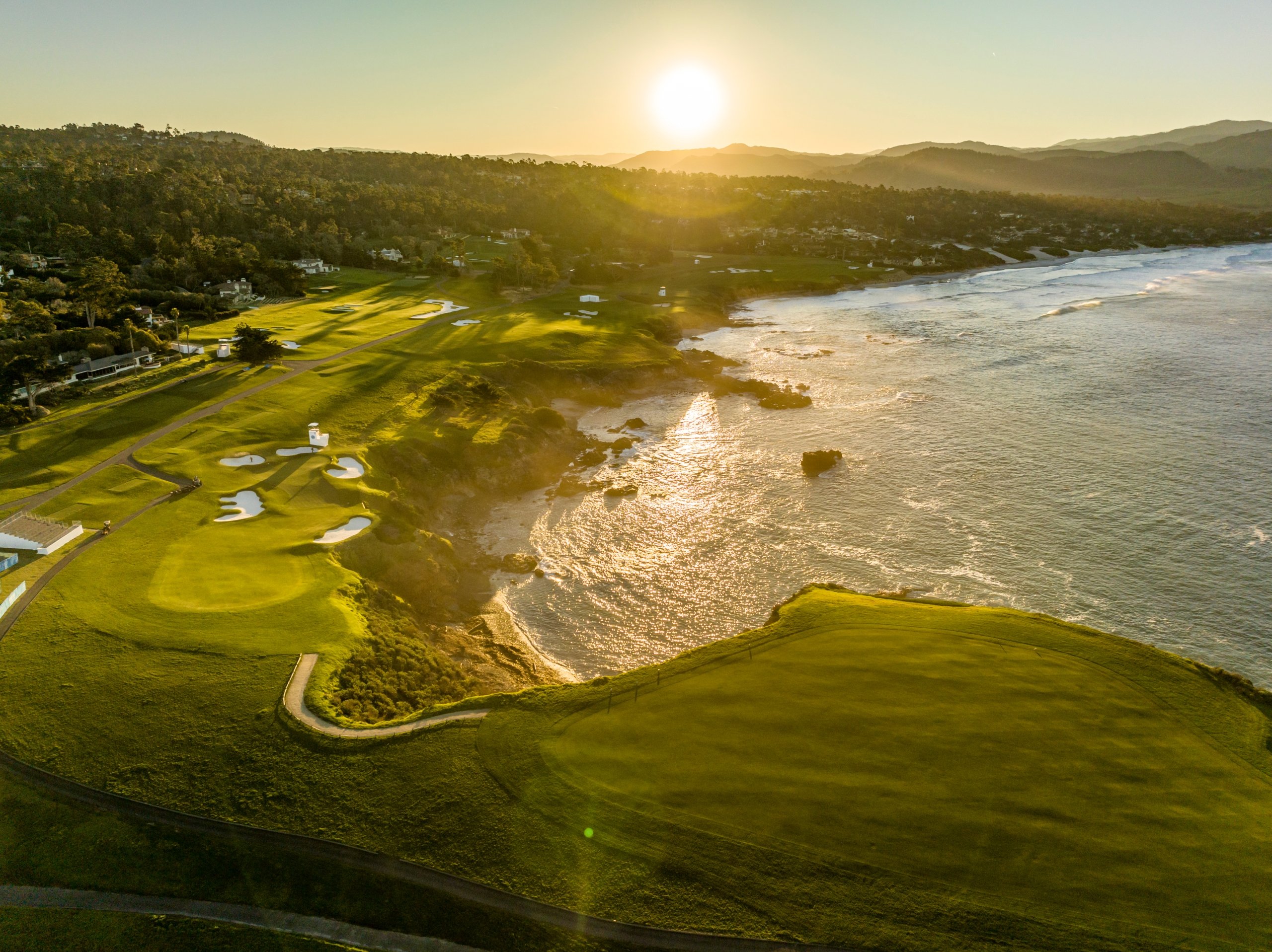 A stunning, high-angle aerial view of a coastal golf course and shoreline bathed in the warm light of the setting sun.