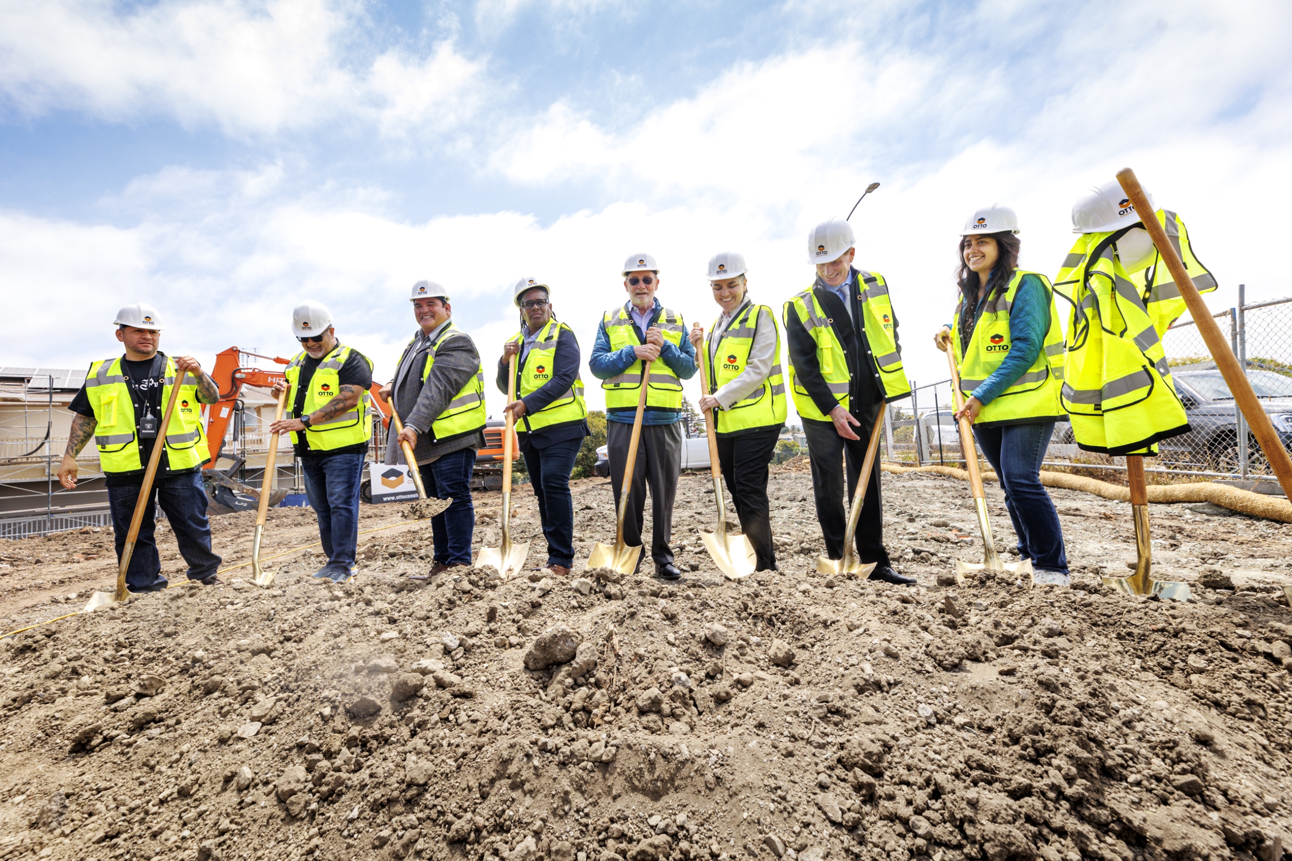 A diverse group of community and construction leaders in hard hats and safety vests holding golden shovels at a groundbreaking ceremony.