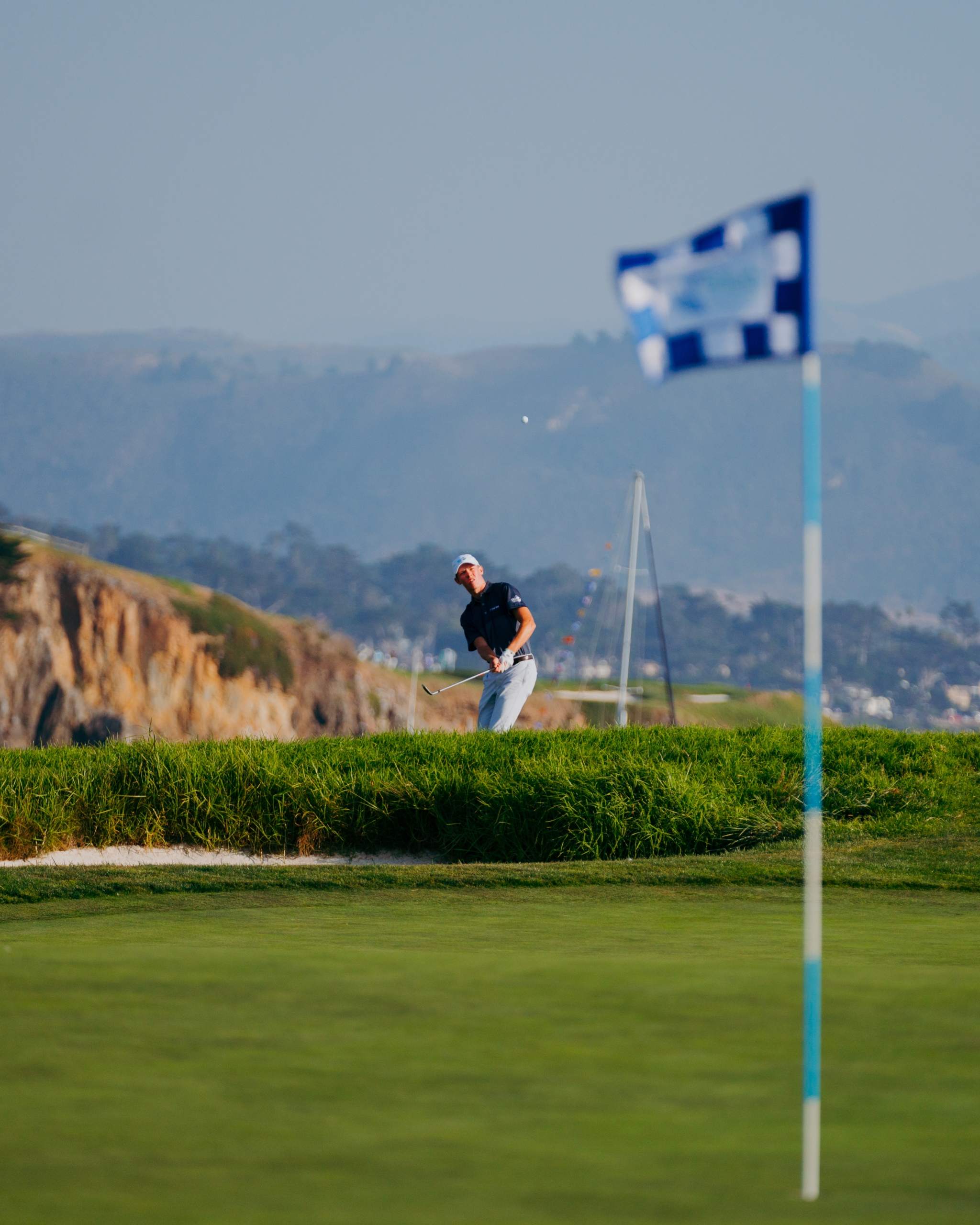 A professional golfer chipping from the rough near a bunker, with the ocean and a checkered flag visible in the background.