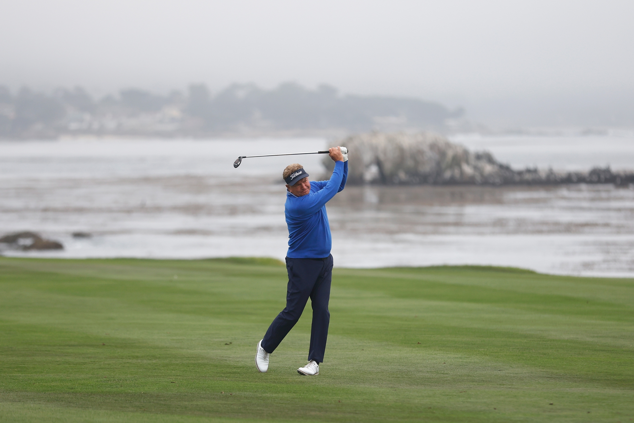 A professional golfer in a blue shirt teeing off on a foggy day on a coastal golf course with the ocean and rocks in the background.