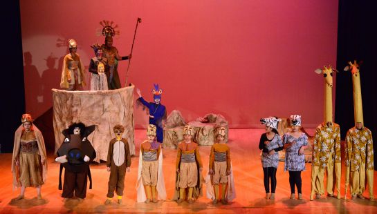 A group of children and adults performing a stage show, possibly a production of The Lion King, in elaborate animal costumes.