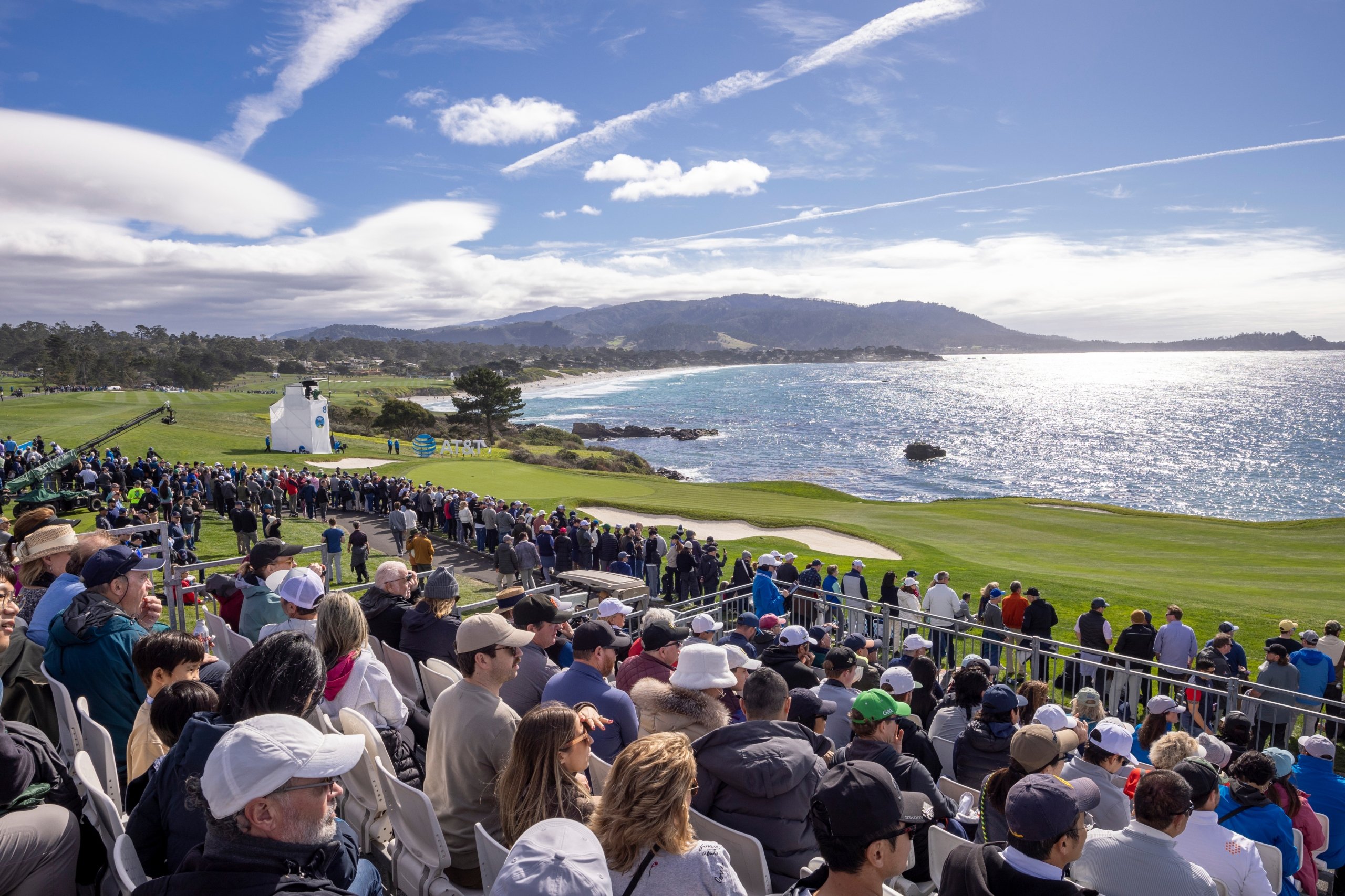 A wide-angle view from the spectator grandstands showing the golf course, the large crowd, and the dramatic coastline under a blue sky.