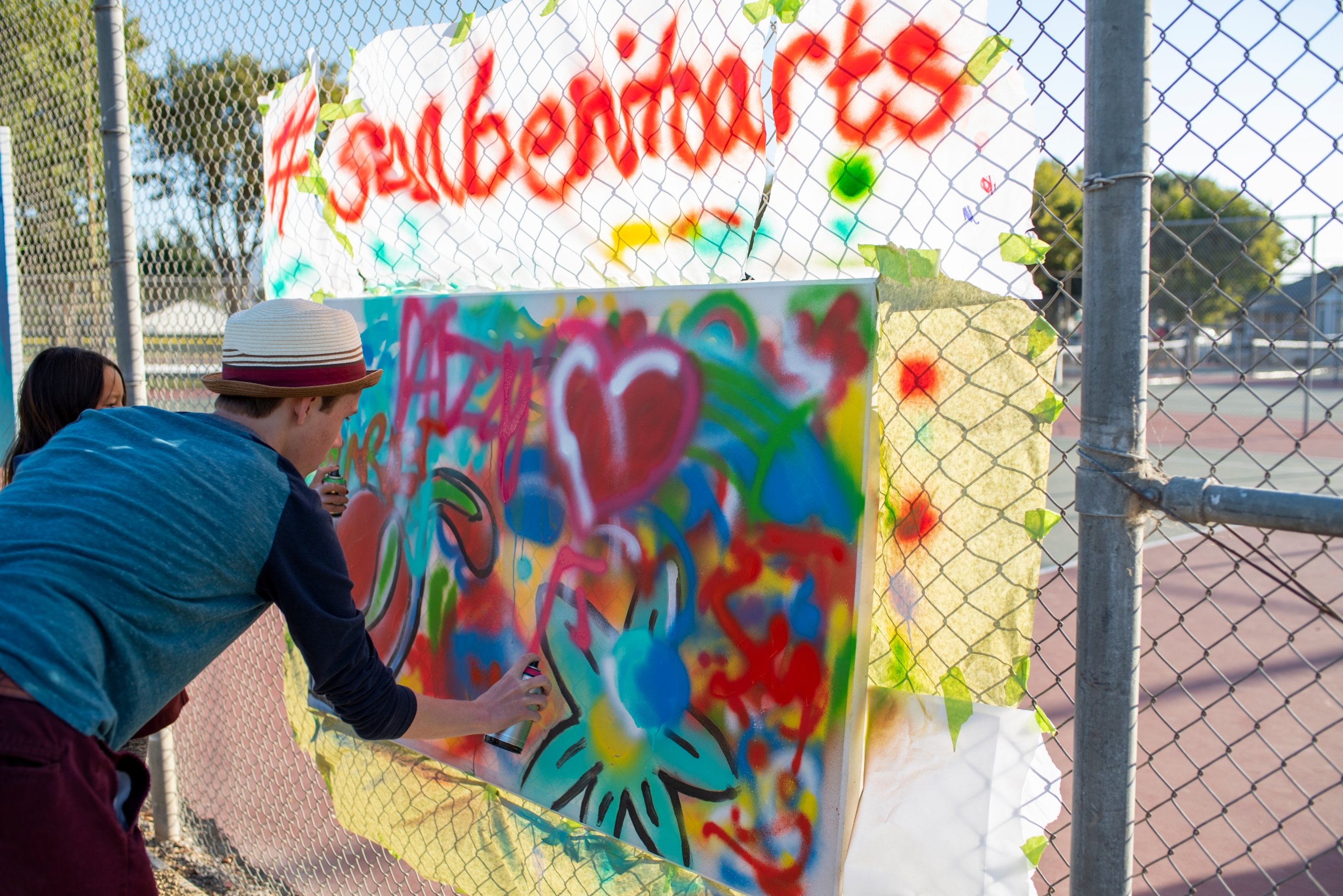 A young person spray-painting a colorful mural on a canvas attached to a chain-link fence, promoting #sanbenitoarts.