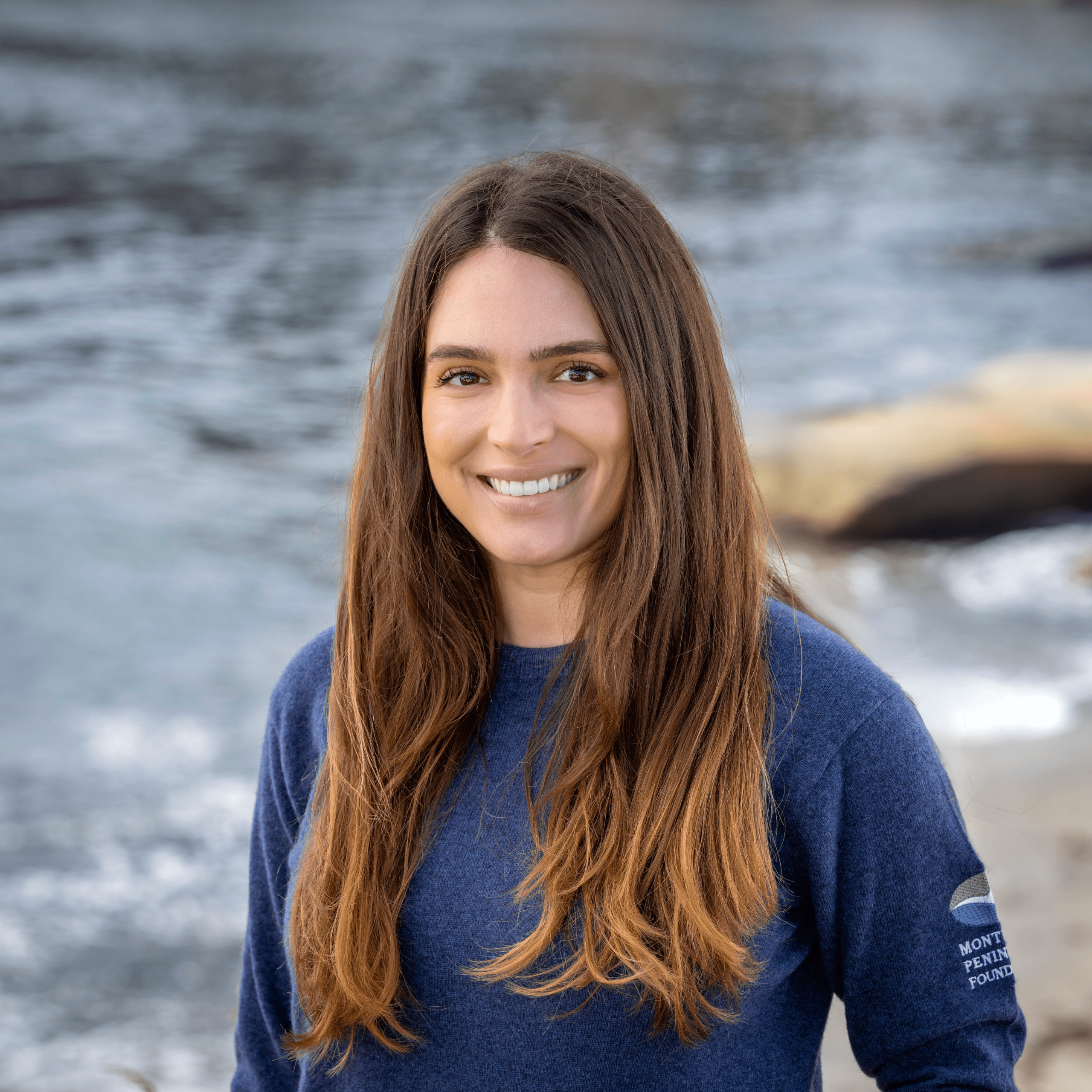 Headshot of a smiling woman with long brown hair wearing a blue MPF branded sweater, standing near the ocean and rocks.