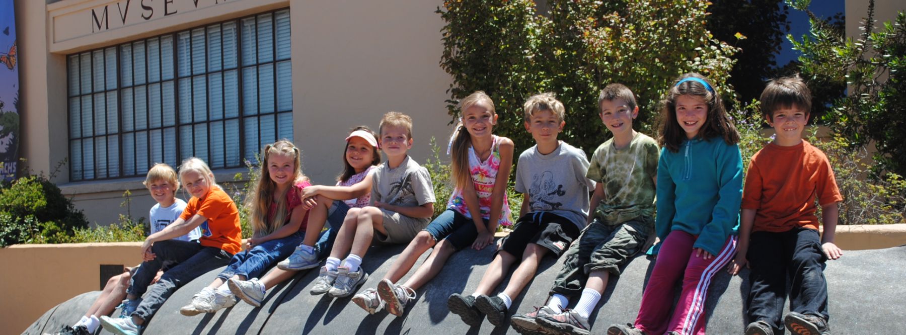 A line of smiling elementary school children sitting outdoors on a ledge in front of the Monterey Museum of Natural History.