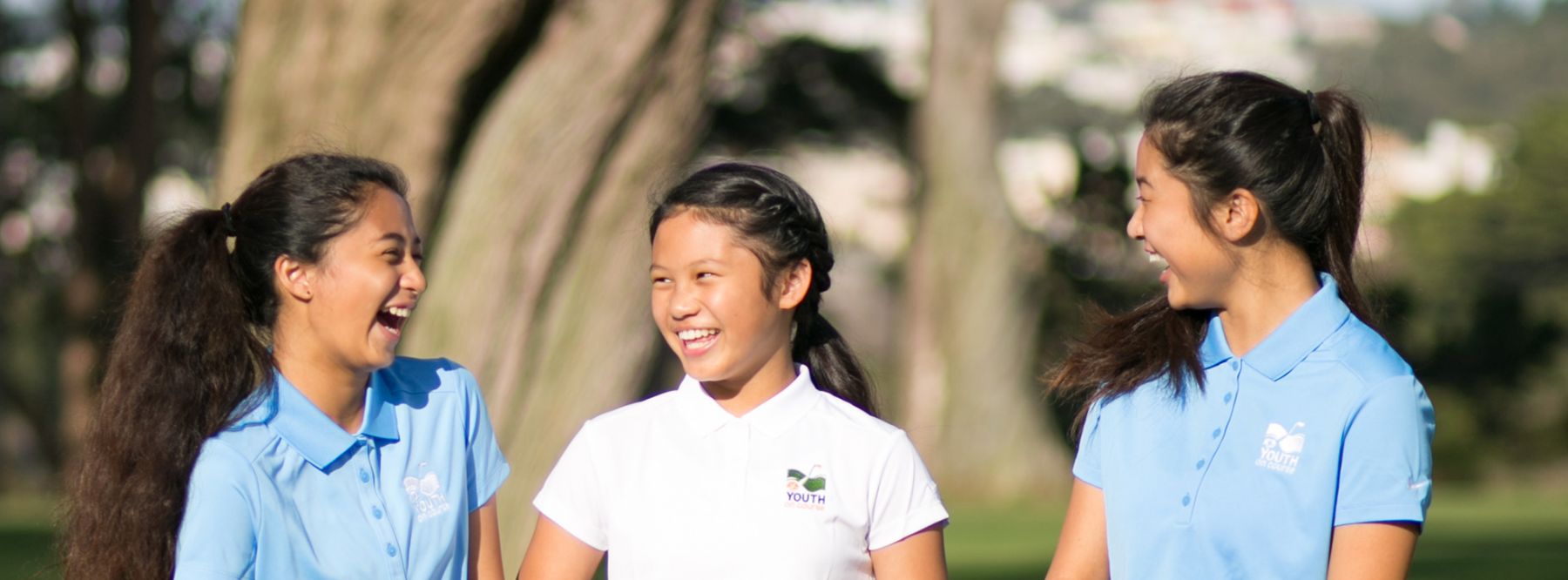 Three young female golfers in Youth on Course polo shirts laughing together outdoors on the golf course.