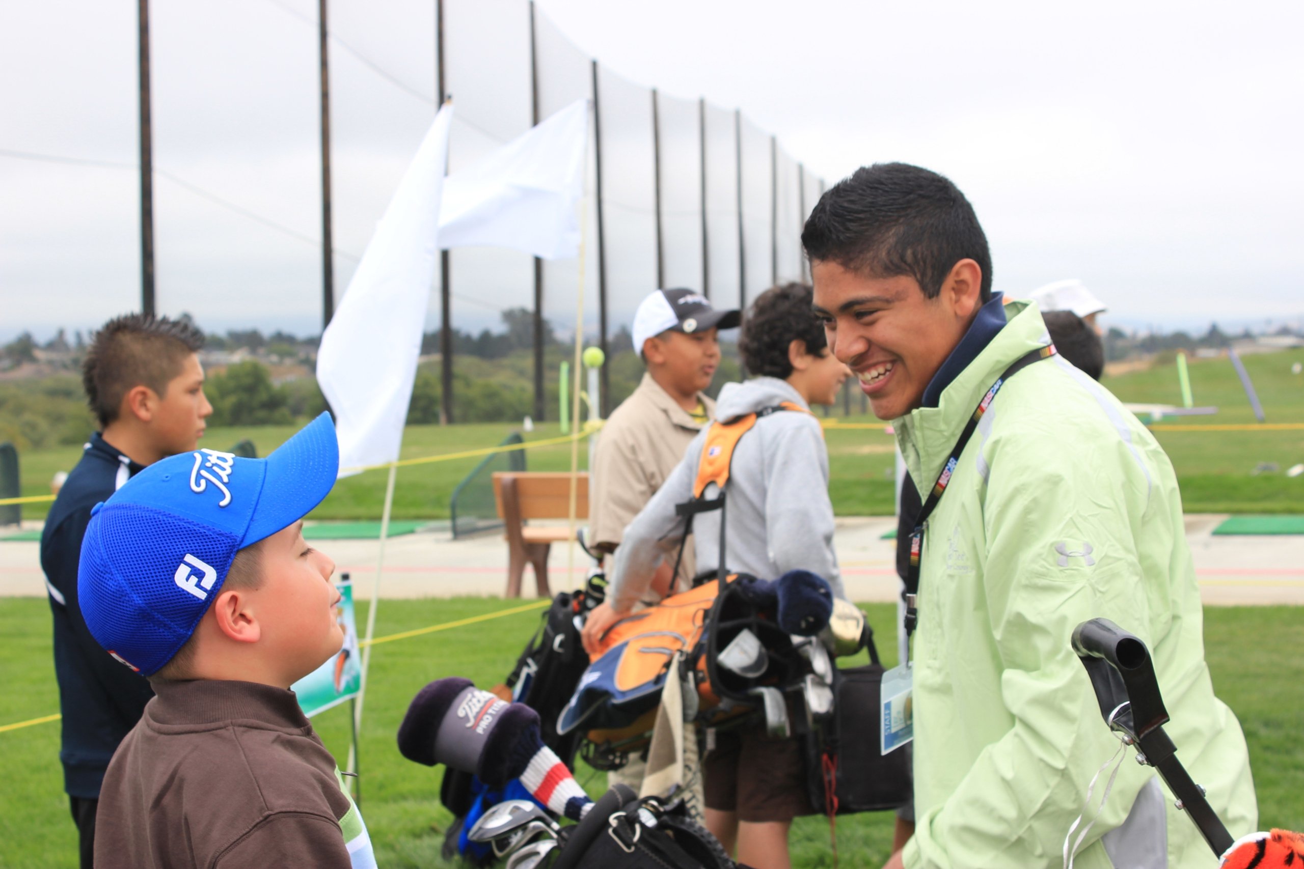 A young boy in a blue golf cap smiles while interacting with an older male golf mentor or youth coach at the driving range.