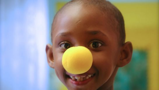 A close-up portrait of a young child smiling while wearing a bright yellow clown nose, representing hospital support programs.