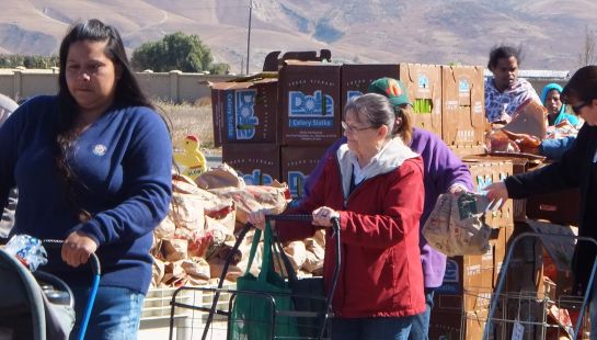 Community members loading groceries into carts and bags at an outdoor food distribution event, with stacked boxes visible.