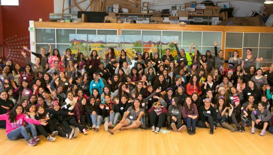 A large group of diverse young women posing and cheering together in a brightly lit community hall.