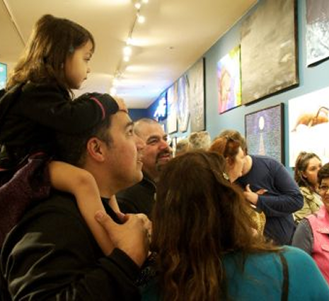 A father holding a young girl on his shoulders while they look at artwork displayed on the wall inside an art gallery or museum.