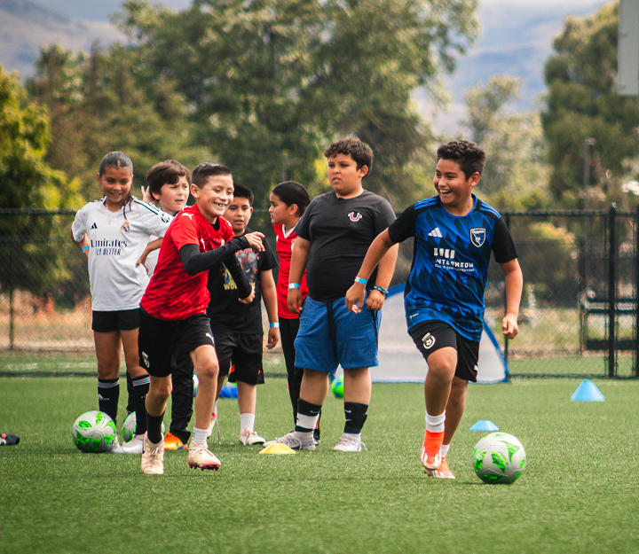 Two boys in soccer jerseys smile while chasing a ball on a green turf field, with other children standing in the background.