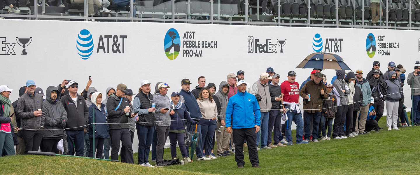 A crowd of spectators watching the tournament from behind a barrier wall branded with AT&T Pebble Beach Pro-Am and FedEx Cup logos.