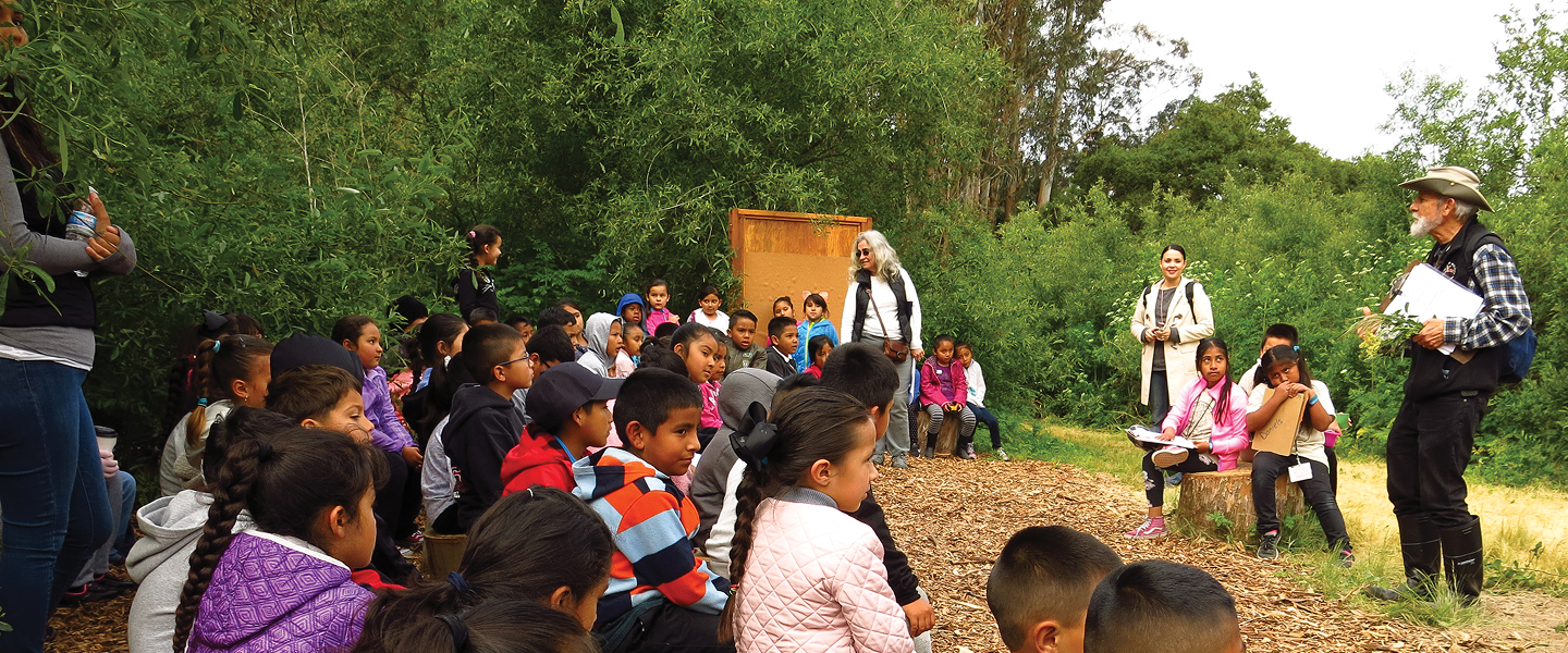 A large group of elementary school students sitting on wood chips during an outdoor environmental education class led by two teachers.