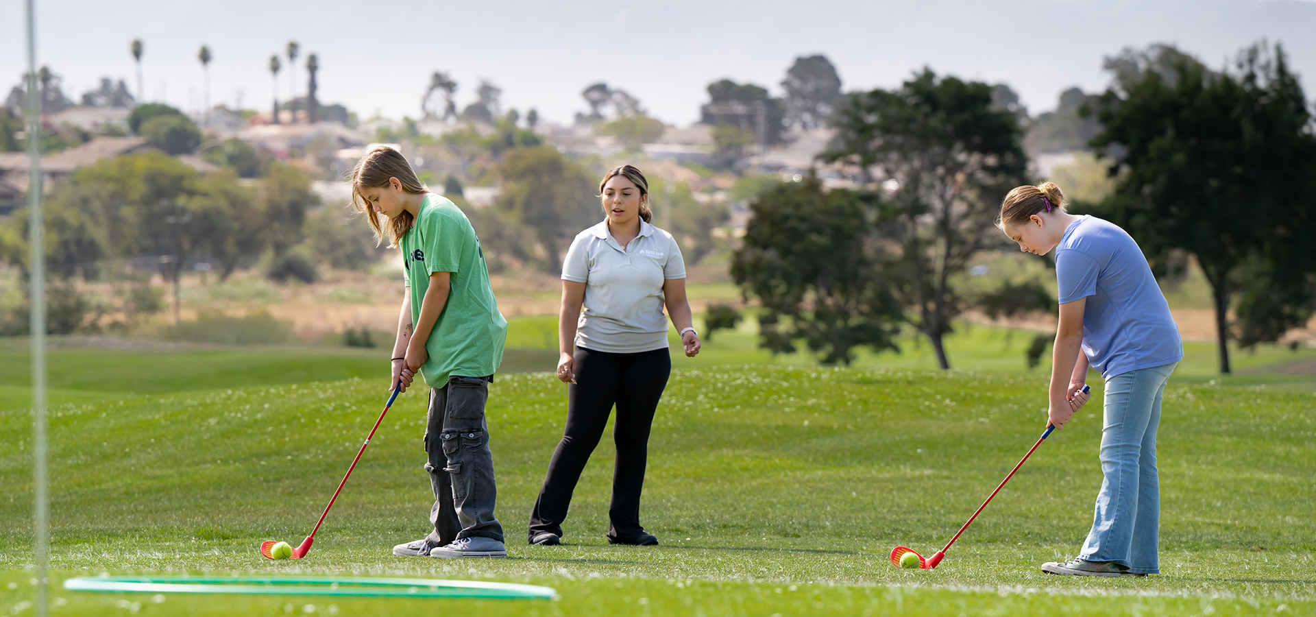 A golf instructor oversees two youth participants practicing their swing on a green field during a First Tee or youth golf clinic.