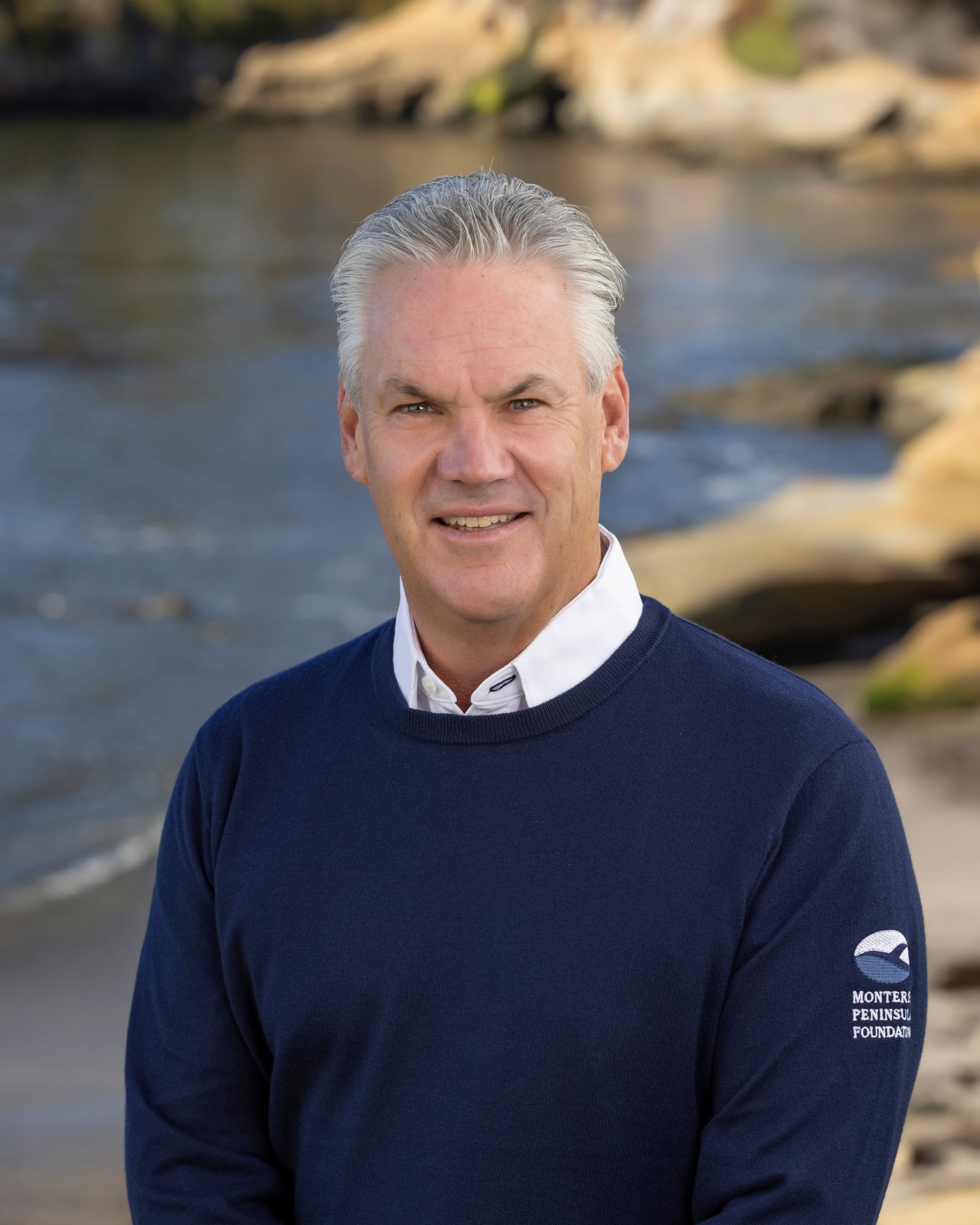 Headshot of Steve John, CEO of the Monterey Peninsula Foundation, smiling while wearing a blue branded sweater, with a coastal background.