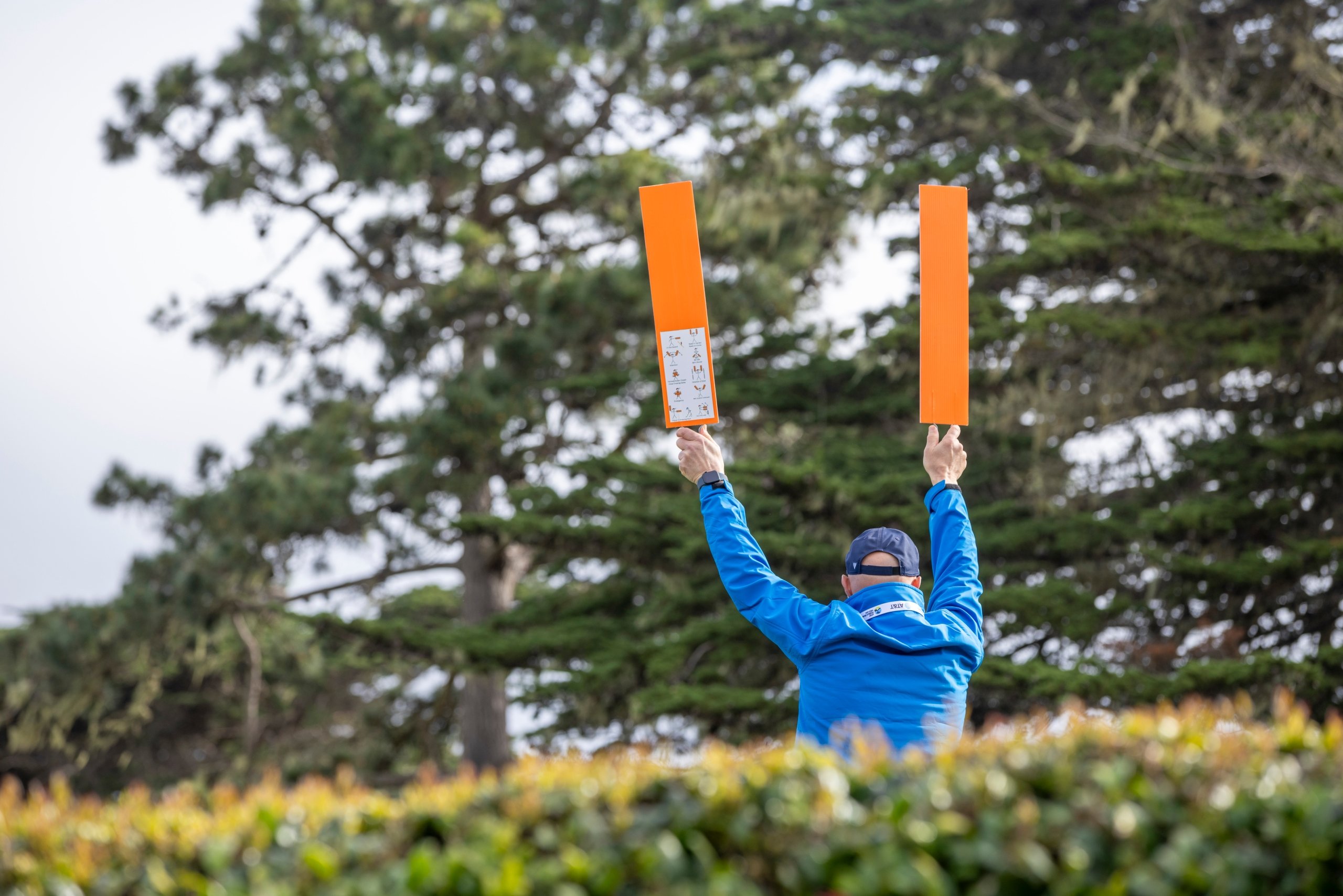 A golf volunteer holding two bright orange penalty indicator boards up in the air against a background of green trees.