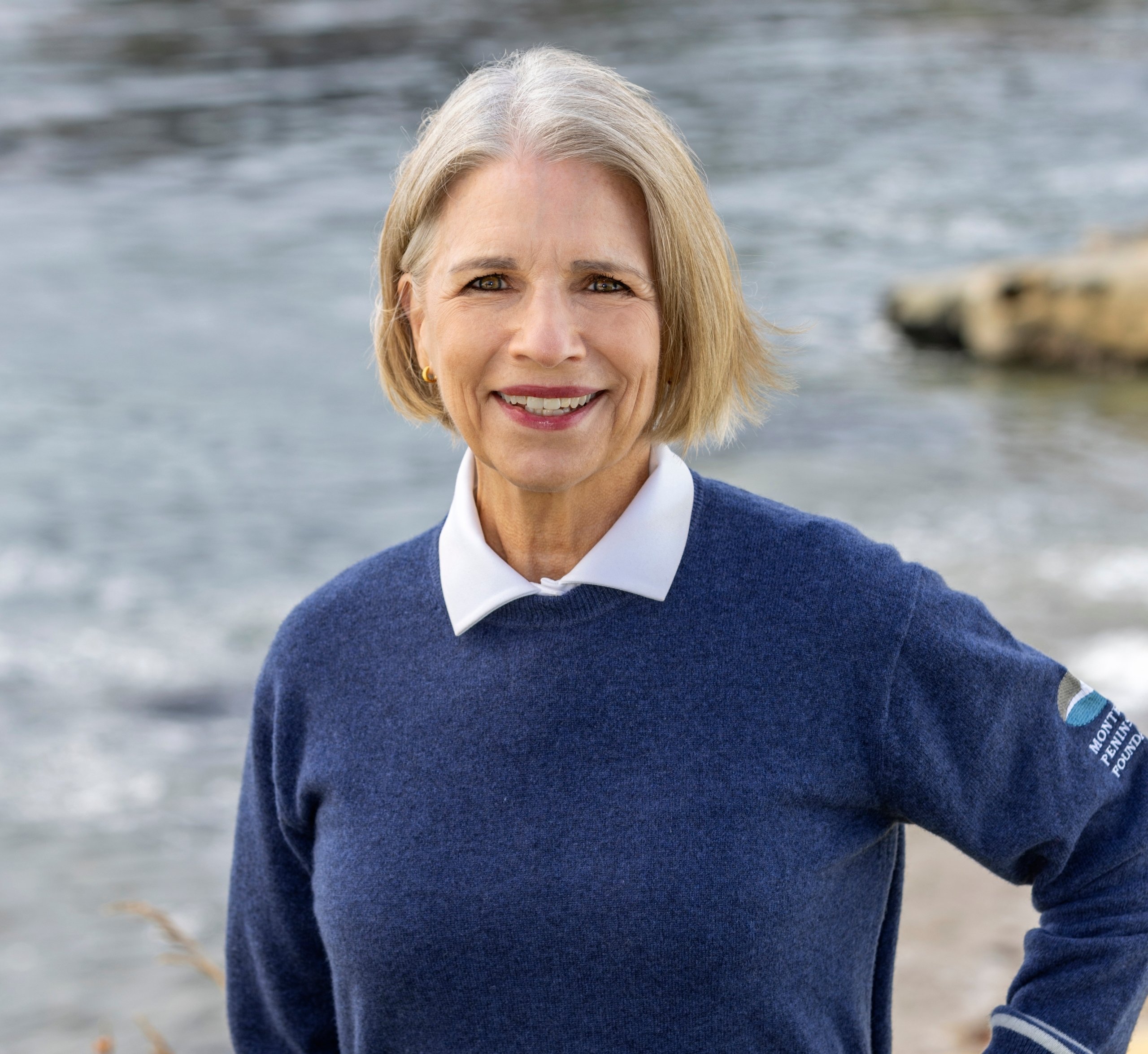 Headshot of a smiling woman with short gray hair wearing a blue sweater over a white collar with the MPF logo, by the shore.
