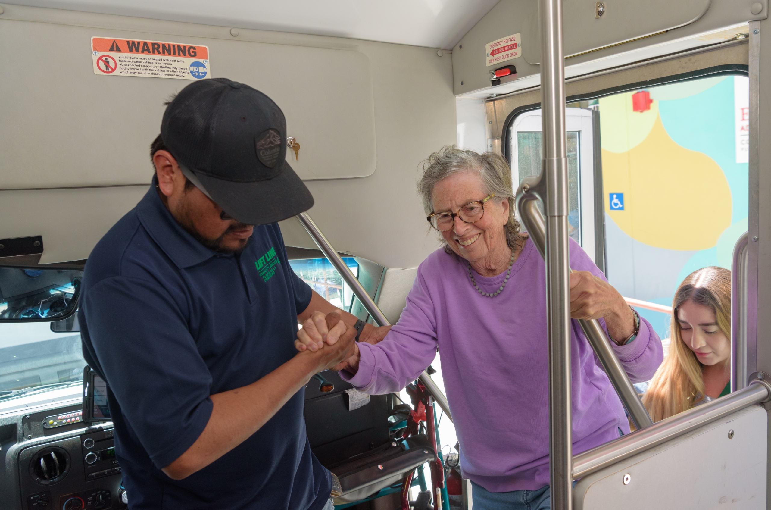 A transit worker assists a smiling elderly woman stepping off a bus, demonstrating accessible community services.