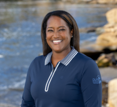 g Headshot of a smiling woman with dark hair wearing a navy blue collared golf shirt, set against a coastal background.