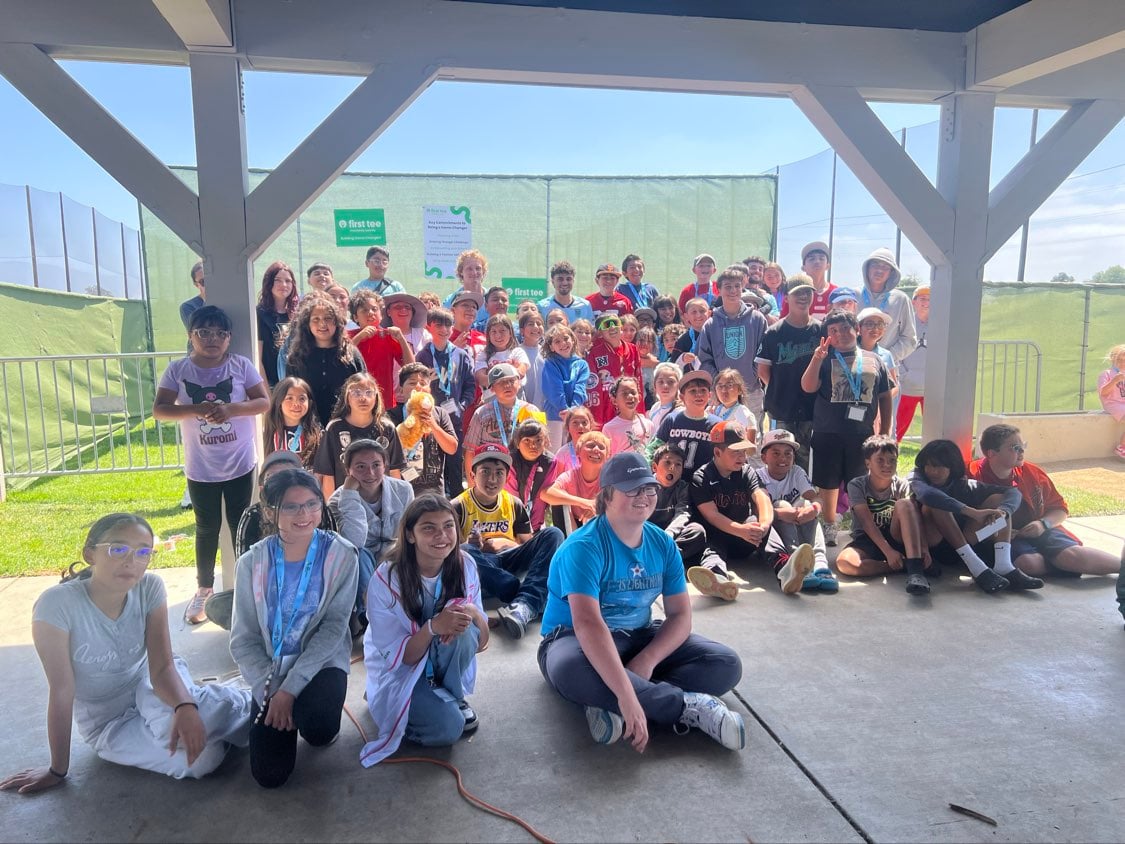 A large group of children and youth sitting and standing together in a sheltered area at a sports camp or clinic.