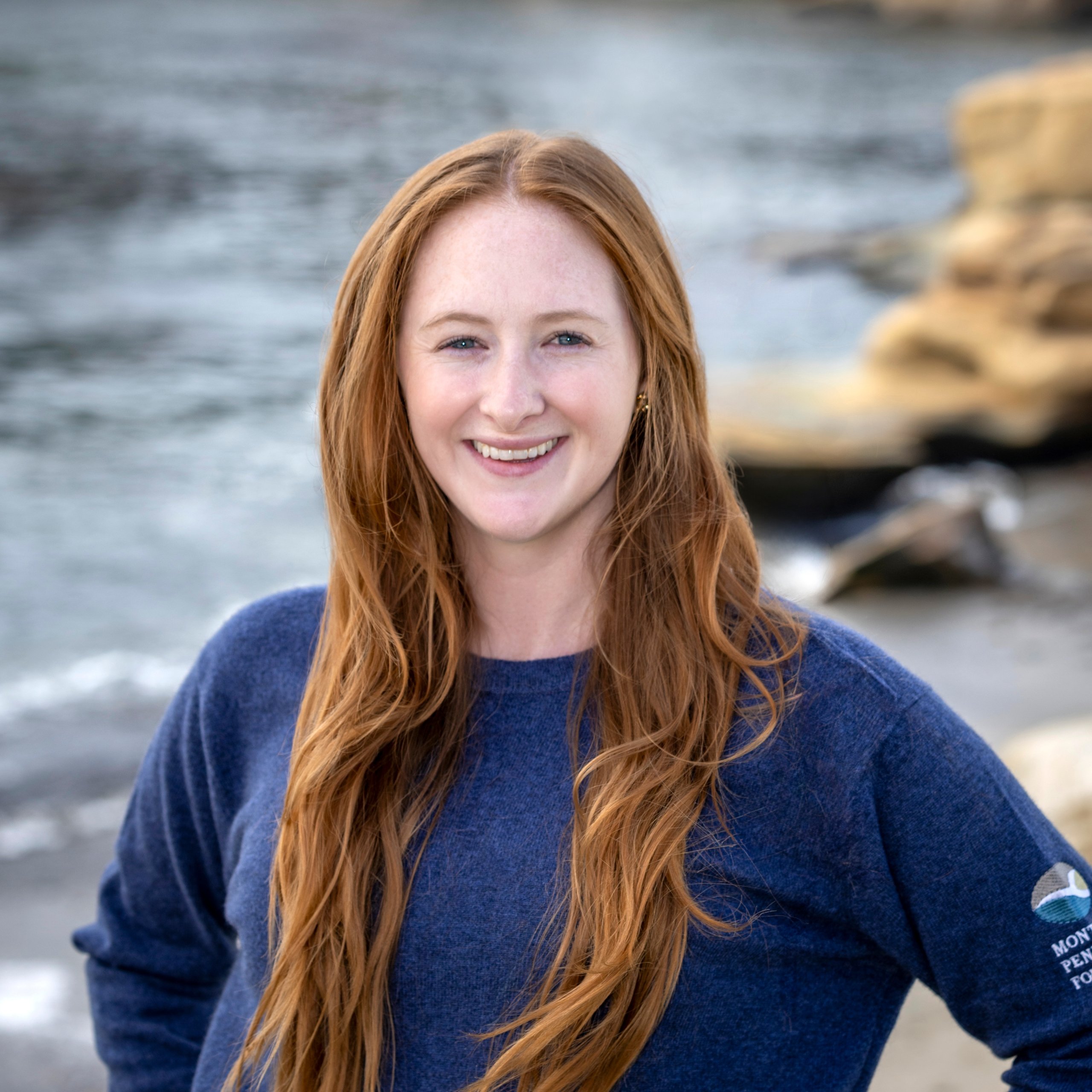 Headshot of a smiling young woman with long red hair wearing a blue MPF branded sweater, standing near the ocean shore.
