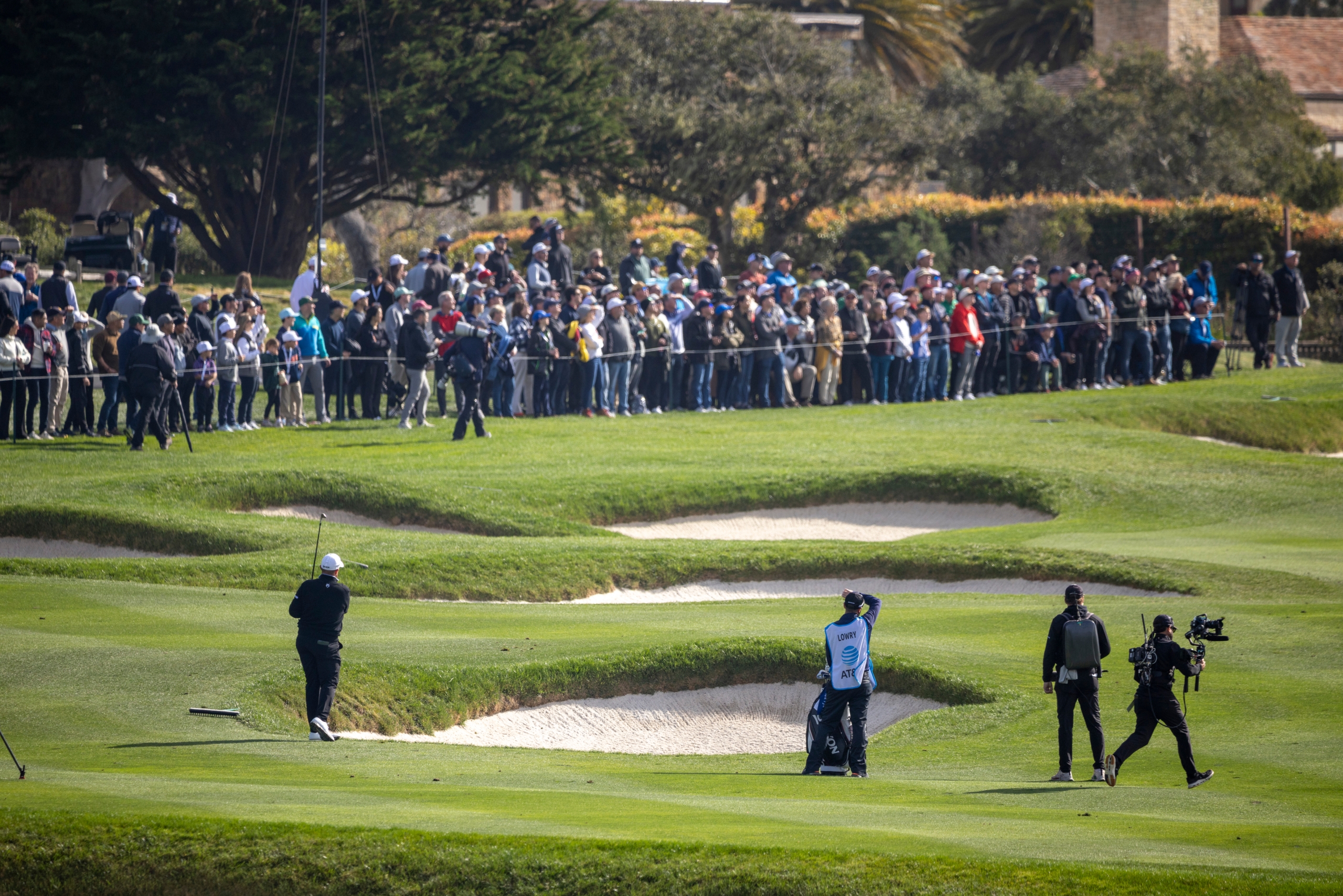 A golfer walking the fairway past several bunkers, followed by cameramen, with a large crowd of spectators watching from the hillside.