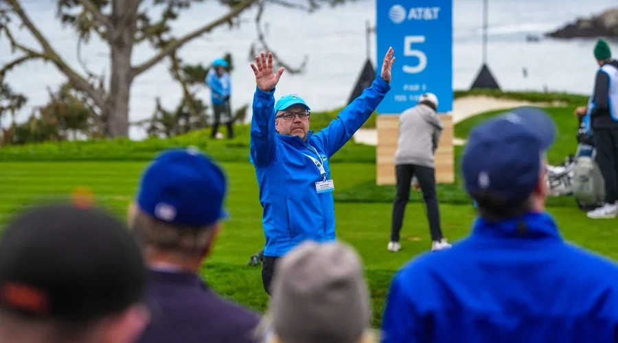 A golf volunteer in a blue jacket raises his arms to quiet the crowd behind the green, with an AT&T scoreboard visible.