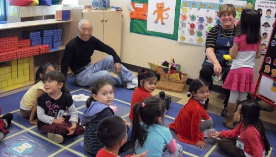 An older man and a woman sitting on the floor in a colorful pre-school classroom surrounded by young children learning.